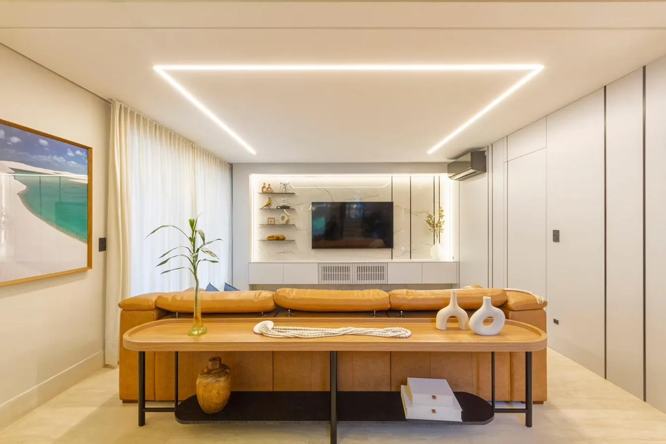Sala de estar de projeto da H+ com painel de porcelanato, sofá de couro caramelo e iluminação linear no teto