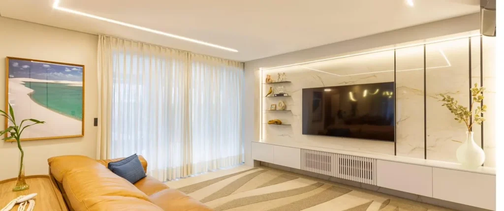 Sala de estar com cortinas em voil, painel de TV em porcelanato e sofá de couro caramelo